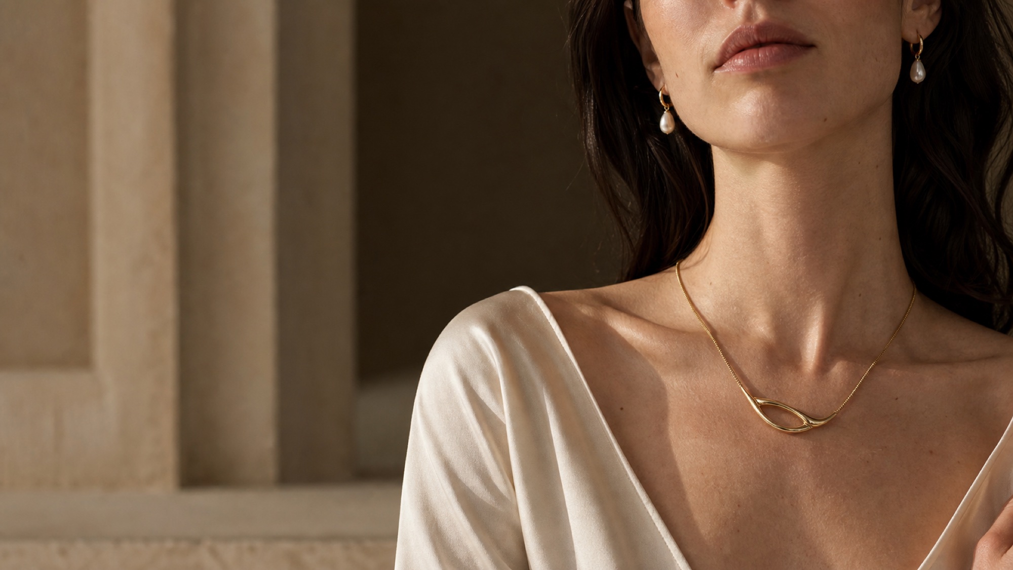 Contemporary gold necklace and pearl earrings styled against warm stone